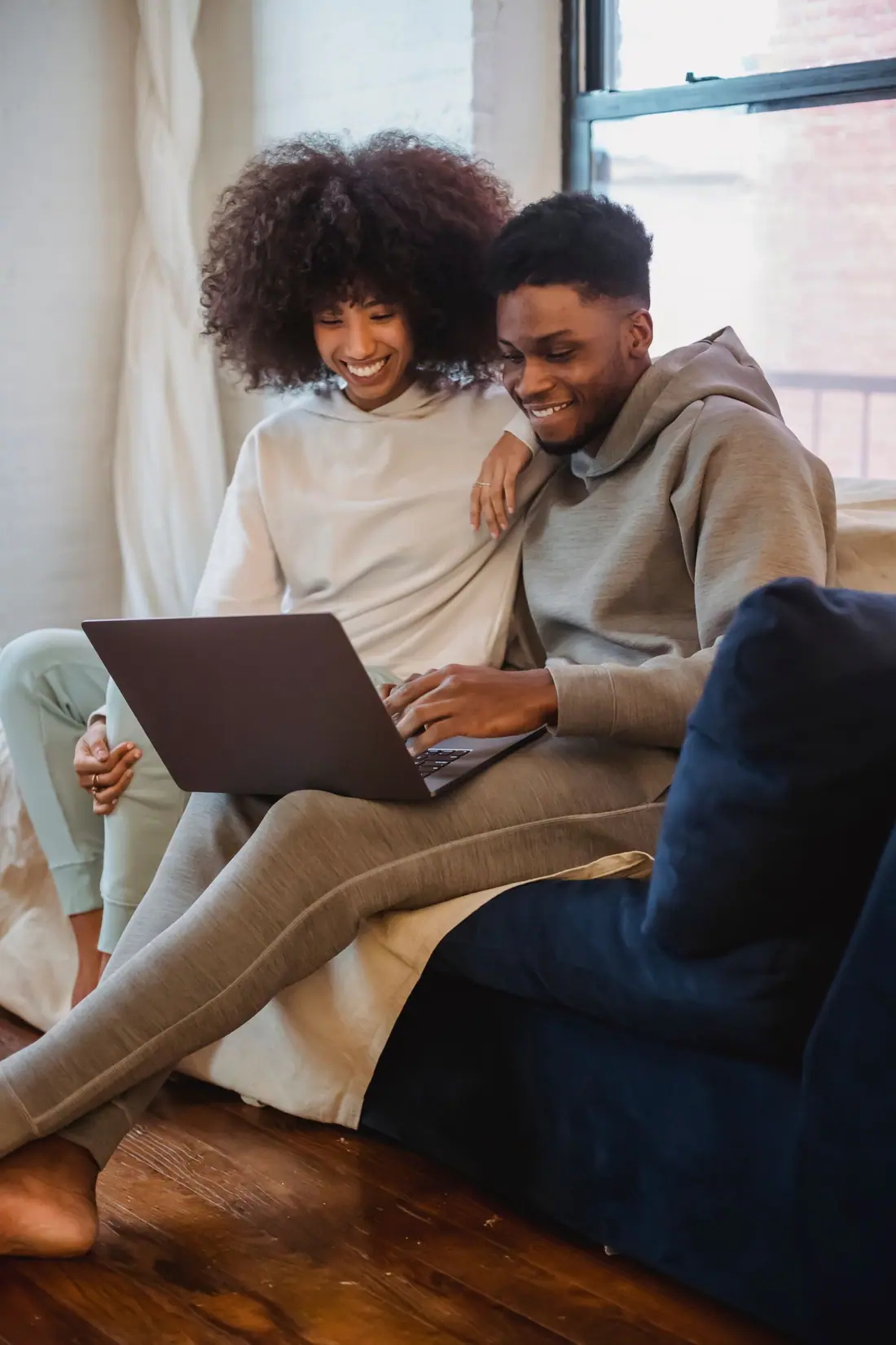 A smiling couple in loungewear sit close on a sofa by a bright window, sharing a laptop