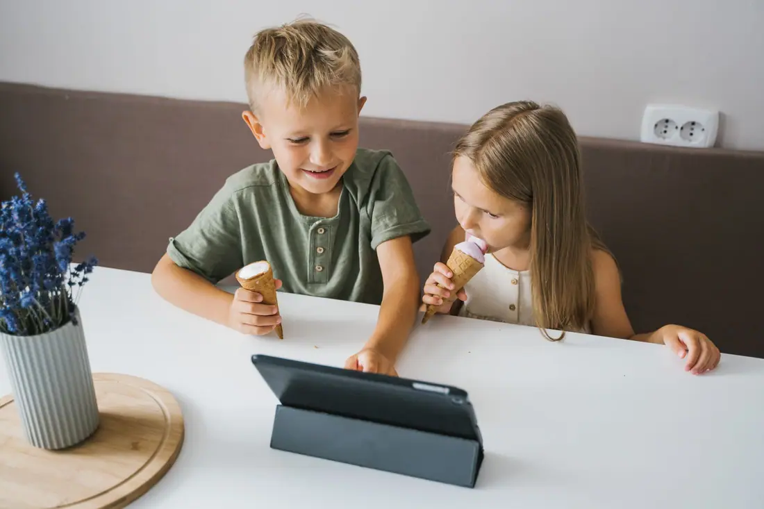 A boy and a girl at a white table smile at a tablet while they eat ice-cream cones