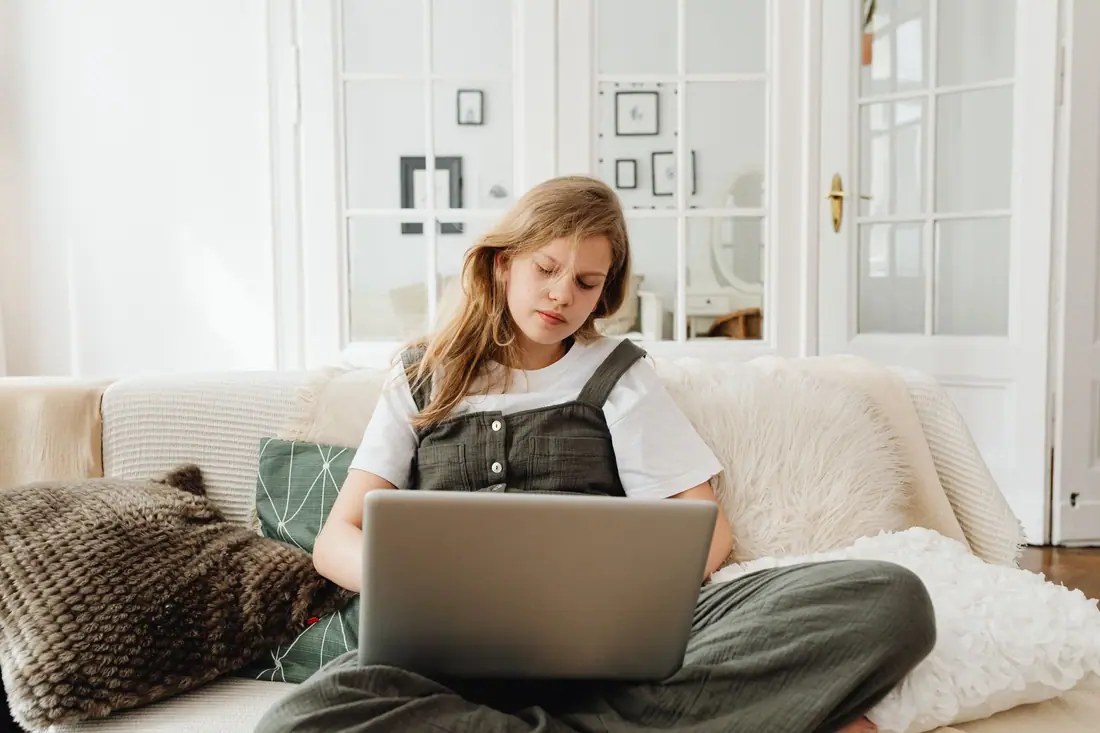 A teenager sits cross-legged on a cream sofa, studying on a laptop