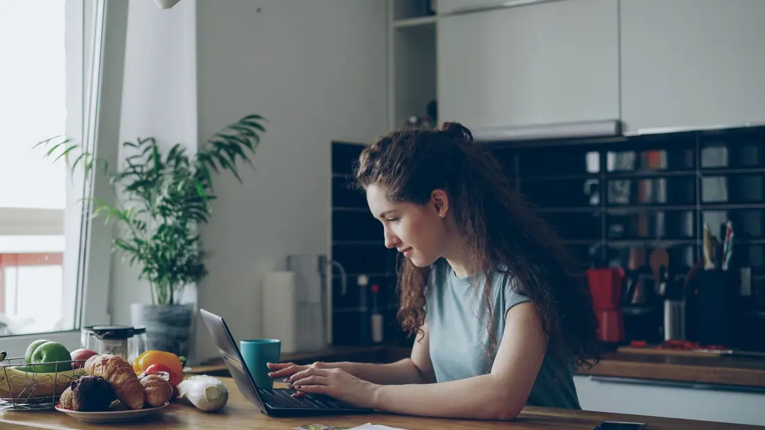 A woman with curly hair types on a laptop at a kitchen table, with a teal mug, croissants and a plant by the window