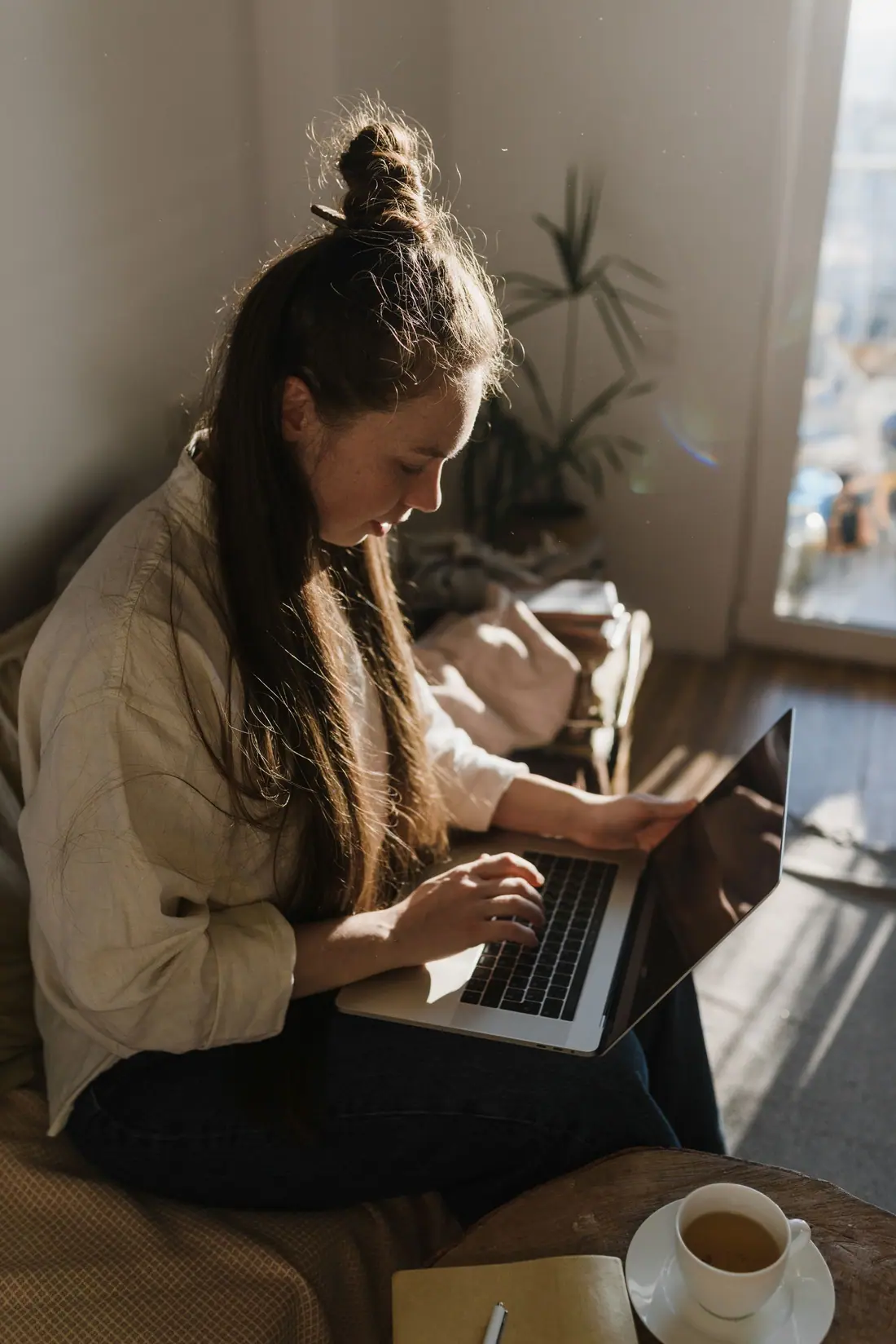 A woman types on a laptop on her lap in a sunlit living room, with a cup of tea beside her