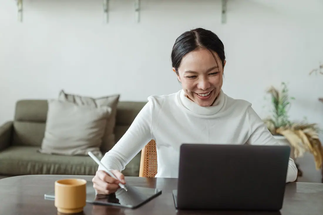 A smiling woman works at a laptop on a home table, with a sofa and a coffee cup nearby